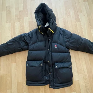 Svart dunjacka från Fjällräven - Säljer en svart dunjacka från Fjällräven med huva och gula snören. Jackan har en dragkedja och knappar framtill samt två stora fickor med knappar. Perfekt för kalla dagar med sin quiltade design.