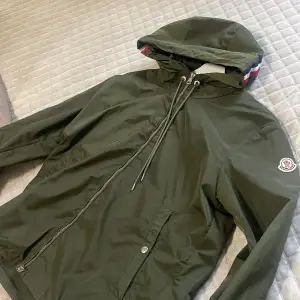 Snygg grön vindjacka från Moncler med huva och dragkedja. Jackan har en stilren design med Moncler-logga på ärmen och snörning vid huvan. Perfekt för blåsiga dagar.