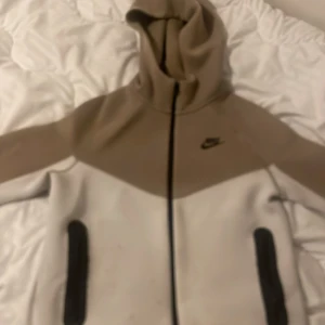 Beige och vit huvtröja från Nike - Säljer en snygg huvtröja från Nike i beige och vit färg. Tröjan har en dragkedja framtill och två praktiska fickor. Perfekt för en avslappnad stil. Huvan ger extra komfort och stil.