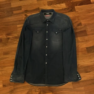 Mörkblå jeansskjorta från Replay - Snygg mörkblå jeansskjorta från Replay med Hyperflex-teknologi. Skjortan har långa ärmar och tryckknappar framtill samt på bröstfickorna. Perfekt för en avslappnad stil med en touch av klass.