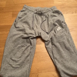 Grå mjukisbyxor från Nike - Säljer ett par grå mjukisbyxor från Nike med elastisk midja och snörning. Byxorna har en avslappnad passform och är perfekta för en chill dag. Nike-loggan är broderad på sidan.