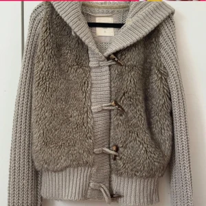 Grå stickad jacka med pälsdetaljer - Säljer en mysig grå stickad jacka med pälsdetaljer framtill. Jackan har långa ärmar och stängs med knappar i trä. Perfekt för kyliga dagar!
