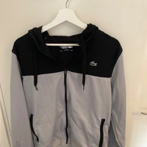 Grå och svart hoodie från Lacoste - Snygg grå och svart hoodie från Lacoste med dragkedja och huva. Den har en broderad krokodil på bröstet och praktiska fickor framtill. Perfekt för en avslappnad stil. Storlek L men passar även M.