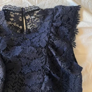 Mörkblå spetsblus från Zara - Snygg mörkblå spetsblus från Zara med blommigt mönster. Blusen är ärmlös och har en elegant design med volangdetaljer. Perfekt för att ge en stilfull touch till din outfit.