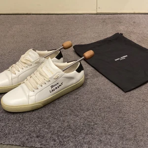 Vita sneakers från Saint Laurent - Snygga vita sneakers från Saint Laurent med svart häl och handskriven logga på sidan. De är välanvända, men med ett bra skick. Skick 7/10. Storlek 43 men sitter som 42. Ändå defekten är insidan av sulan, men inget som märks vid användning utan bara utseende på insidasulan. Priset kan diskuteras.