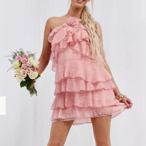 Rosa axelbandslös klänning med volanger - Charmig rosa axelbandslös klänning med flera lager av volanger och en dekorativ blomma vid halsen. Perfekt för somriga tillfällen med sin luftiga och lekfulla design. Den är helt oanvänd!! Pris lappen sitter kvar 