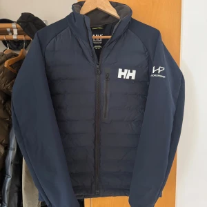 Mörkblå jacka från Helly Hansen - Säljer en mörkblå jacka från Helly Hansen med dragkedja och logotyp på bröstet och ryggen. Jackan har långa ärmar och är perfekt för kyligare dagar. Den är designad för att hålla dig varm och bekväm. Bara att höra av sig vid fler bilder och frågor