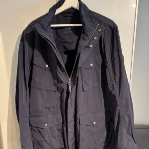 Blå jacka från Hackett London - Snygg svart jacka från Hackett med dragkedja och knappar. Jackan har flera praktiska fickor och en stilren design. Perfekt för höst och vår. Nypris 5000.