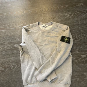 Grå sweatshirt från Stone Island - Säljer en stilren grå sweatshirt från Stone Island med deras ikoniska märke på ärmen. Tröjan har en klassisk rund hals och långa ärmar, perfekt för en avslappnad look. Passar bra till både jeans och joggers.