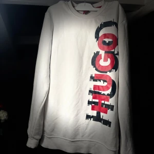 Vit tröja från Hugo - Säljer en vit tröja från Hugo med ett stort tryck i rött och svart på framsidan. Tröjan har långa ärmar och en rund halsringning. Perfekt för en avslappnad stil.