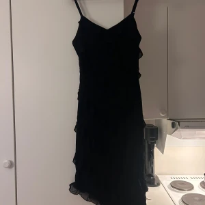 Svart klänning med volangdetaljer - Snygg svart klänning med smala axelband och volangdetaljer. Perfekt för en stilren look. Klänningen har en elegant passform och är tillverkad i ett lätt material. Storlek S, säljer då den ej passar