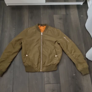 Olivgrön bomberjacka med orange foder. - Säljer en snygg olivgrön bomberjacka med orange foder. Jackan har dragkedja framtill och ribbade muddar vid ärmslut och nederkant. Perfekt för en cool och avslappnad stil.