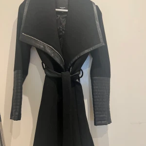 Svart kappa med quiltade detaljer - Snygg svart kappa i ull med quiltade detaljer på ärmarna och en bred krage med dragkedja. Jackan har ett bälte i midjan för en figursydd look. Från märket Rockandblue