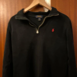 Svart tröja med dragkedja från Ralph Lauren - Snygg svart tröja från Ralph Lauren med en liten röd broderad logotyp på bröstet. Tröjan har en dragkedja vid halsen och långa ärmar. Perfekt för en stilren look.