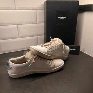 Vita sneakers från Saint Laurent - Snygga vita sneakers från Saint Laurent med unika läppdetaljer i rött och blått på hälen. Model sl/01, perfekta inför sommaren. Finns en del skavanker och dem är rätt så välanvända. Box, påse och certifikat med följer, inne sulan är dock inte orginal. Hör av er vid minska fråga✌️