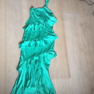 Grön långklänning från Boohoo - Säljer en snygg grön långklänning från Boohoo med volanger som ger en härlig rörelse. Klänningen har en axelbandslös design och är perfekt för sommarens festligheter. Materialet är mjukt och glänsande, vilket ger en lyxig känsla.