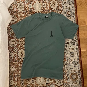 Grön vintage Stussy t-shirt  - Snygg grön t-shirt från Stussy med ett coolt tryck av en figur på både fram- och baksidan. T-shirten är kortärmad och tillverkad i bomull, perfekt för en avslappnad stil. Också perfekt till sommaren.