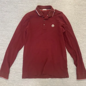 Vinröd pikétröja från Moncler - Snygg vinröd pikétröja från Moncler med långa ärmar och klassisk krage. Tröjan har knappar framtill och Monclers logga på bröstet. Kragen har en stilren randig detalj i vitt och rött.