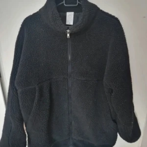 Svart teddyjacka med dragkedja - Mysig svart teddyjacka med dragkedja och hög krage. Perfekt för kyliga dagar när du vill hålla dig varm och bekväm. Jackan har en avslappnad passform och långa ärmar.