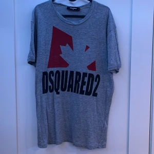 Dsquared2 T shirt  - Säljer en grå t-shirt från Dsquared2 med ett stort tryck av en röd lönnlöv och texten 'DSQUARED2' i svart på framsidan. T-shirten har en rund hals och korta ärmar. Perfekt för en avslappnad stil. ”Pris kan diskuteras!”