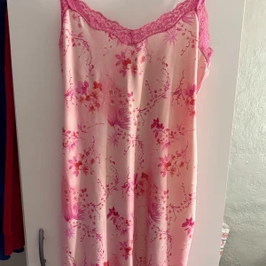 Rosa blommig klänning med spetsdetaljer - Charmig rosa klänning med blommigt mönster och spetsdetaljer vid halsringningen. Klänningen har smala axelband och en söt liten rosett. Perfekt för en somrig look!