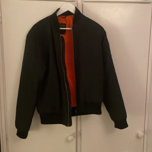 As cool bomberjacka - Snygg svart bomberjacka med ett coolt orange foder. Jackan har en dragkedja framtill och en extra dragkedja på ärmen för en edgy look. Perfekt för att ge din outfit en stilren touch.