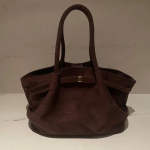 Brun mocka handväska - Snygg brun handväska i mjuk mocka med två handtag. Väskan har en stilren design med en liten gulddetalj framtill. Aldrig använd 