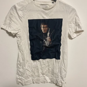 Vit t-shirt från Limitato - Säljer en vit t-shirt från Limitato med ett tryck av James Bond på framsidan. T-shirten är kortärmad och har en klassisk passform. Perfekt för dig som gillar unika tryck och stilren design.