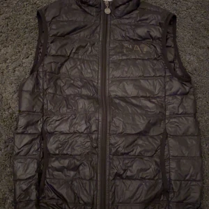 Svart dunväst från Emporio Armani - Snygg svart dunväst från Emporio Armani med dragkedja framtill. Västen har en quiltad design och är perfekt för kyligare dagar. Märkesloggan EA7 syns tydligt på bröstet. Ett stilrent val för den modeintresserade.