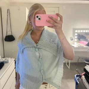 Ljusblå stickad kofta med pärldetalj - Superfin ljusblå stickad kofta med stora ribbade krage och knappar framtill. Koftan har en unik pärlbroderad detalj på kragen och är lite loose i modellen med kortare ärmar. Perfekt att slänga över en topp för extra mysig stil.