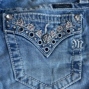 Miss Me caprijeans med detaljer, stl 26 - Snygga blå caprijeans från Miss Me med grymma detaljer på bakfickorna som nitar, broderier och bling. Jeansen har slitningar och en cool tvättad look. Perfekta för dig som gillar statement-jeans med extra attityd.