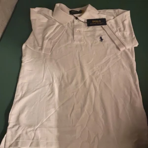 Vit pikétröja från Polo Ralph Lauren - Vit pikétröja från Polo Ralph Lauren med klassisk krage, korta ärmar och broderad marinblå logga på bröstet. Tröjan är i bomull och har en clean, tidlös look som passar perfekt till jeans eller shorts.