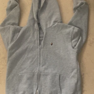 Grå hoodie från A Bathing Ape XL - Snygg grå hoodie från A Bathing Ape i storlek XL. Tröjan har dragkedja, huva och klassisk BAPE-logga på bröstet. Tillverkad i mjuk bomull och har praktiska fickor framtill. Perfekt för en chill streetwear-look.
