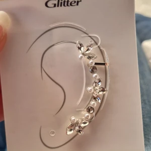Glittrig ear cuff med pärlor  - Snygg ear cuff i silverfärg med glittriga stenar och vita pärlor. Bladformade detaljer ger ett elegant och trendigt intryck. Perfekt för dig som vill sticka ut med ett unikt örhänge som klättrar längs örat.