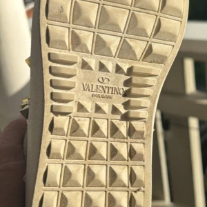 Valentino Rockstud sneakers vit/rosa - Snygga Valentino Rockstud sneakers i vitt skinn med ljusrosa detaljer och ikoniska guldiga nitar. Skorna har snörning, platt sula och rund tå. Perfekta för dig som vill sticka ut med en lyxig och edgy vibe.