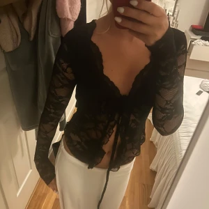 Svart spetsig blus med knyt - Säljer en svart blus i transparent spets med långa ärmar och v-ringning. Toppen har knyt framtill och är croppad, aldrig använd 