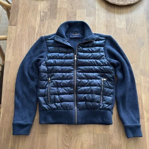 Mörkblå cardigan från Vonroe - Snygg mörkblå jacka från Vonroe med quiltad, pufferliknande framsida och stickade ärmar samt rygg. Jackan har dragkedja framtill, två fickor med dragkedja och hög krage. Nypris är 1000kr.