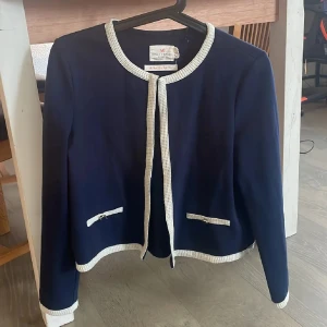 Mörkblå kavaj från Holly & White - Snygg mörkblå kavaj med vita detaljer från Holly & White. Rak passform, rund halsringning och dekorativa fickor framtill. Kavajen har vita ribbade kanter längs krage, ärmslut och nederkant för en klassisk look. Perfekt för dig som gillar stilrena plagg.