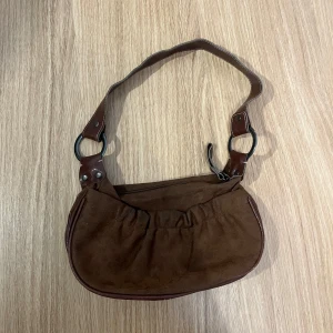 Brun mocka axelväska med ringdetaljer - Säljer en brun axelväska i mocka med snygga metallringar och detaljer i brunt skinn. Väskan har en mjuk, rundad form och dragkedja upptill. Perfekt storlek för det viktigaste och har en cool vintagevibe. Väldigt älskad väska i ok skick.