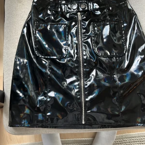 Svart glansig kjol från River Island - Snygg svart kortkjol från River Island i glansigt syntetmaterial. Kjolen har två stora fickor framtill och en silverfärgad dragkedja som går hela vägen upp. Perfekt för dig som gillar statement-plagg med cool vibe. Har bara använt en gång.