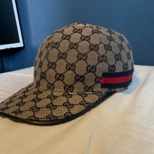 Gucci keps med GG-mönster - SKICKA PRISFÖRSLAG Snygg keps från Gucci i beige med klassiskt GG-monogram över hela kepsen. På sidorna finns Guccis ikoniska röd-blå rand. Kepsen är tillverkad i bomull och har en böjd skärm. Perfekt för dig som vill sticka ut med en lyxig accessoar.