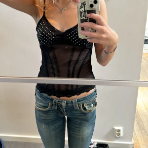 Svart korsett-topp med nitar och spets - Supersnygg svart korsett-topp med smala axelband, dekorerad med silvriga nitar över bysten och spetskant nertill. Toppens tyg har markerade sömmar som ger en cool form och detaljerad look. Perfekt för dig som vill sticka ut med en edgy vibe.