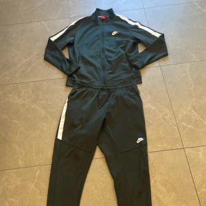 Svart Nike träningsväst med vita ränder - Snygg svart träningsväst från Nike med vita ränder längs axlarna. Västen har dragkedja framtill, ståkrage och klassisk Nike-logga på bröstet. Materialet är mjukt och stretchigt, perfekt för sportiga outfits eller chill dagar.