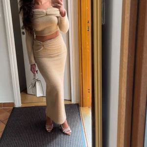 Beige set med twistdetalj - Supersnygg beige långkjol och off shoulder tröja med twistdetalj framtill i midjan och axeln. Kjolen är figurnära och har en mjuk, stickad känsla som ger en chill men ändå elegant vibe. Perfekt att matcha med crop top eller stickad tröja för en trendig look.