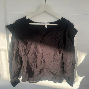 Svart blus med knappar från H&M - Svart blus från H&M med v-ringning och knappar framtill. Långa ärmar och lätt puff vid axlarna ger en cool siluett. Perfekt att styla med jeans eller kjol för en trendig look.