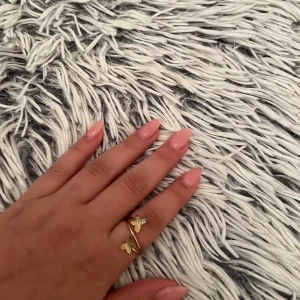Guldfärgad ring med fjärilar - Snygg guldfärgad ring med två detaljerade fjärilar som sitter på en tunn, öppen ring. Fjärilarna har en glittrig yta som fångar ljuset och ger en trendig look. Perfekt accessoar för dig som gillar unika smycken med lite extra bling.