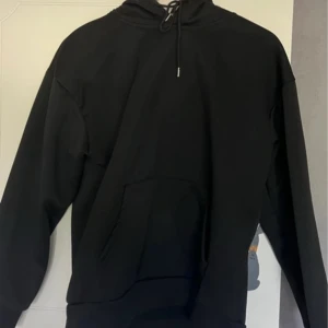 Svart hoodie med huva och ficka - En klassisk svart hoodie med huva och snörning, samt en stor magficka framtill. Tröjan har lång ärm och en relaxed passform, perfekt för chill dagar eller när du vill ha en enkel och stilren look.