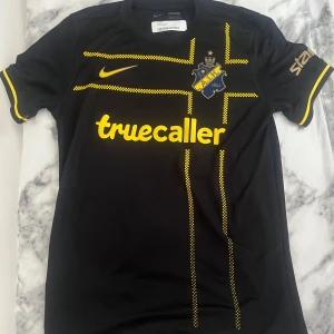 AIK fotbollströja Nike svart/gul M - AIK:s officiella fotbollströja från Nike i svart med gula detaljer och rutmönstrade linjer. Tröjan har klubbmärke på bröstet och Truecaller-logga. Köpt för 1300kr, aldrig använt!