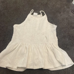 Vit volangtop från Zara - Supersöt vit topp från Zara med smockad överdel och volang nertill. Tunna axelband och luftig passform gör den perfekt för varma dagar. Enkel och stilren design som passar till jeans eller kjol.