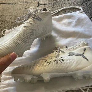 Fotbollsskor signerade av Carolina seger - Säljer ett par vita Puma fotbollsskor med grå detaljer och snörning. Skorna har stickad ovandel och platt sula med dobbar för gräsplan. Snyggt mönstrade skosnören och Puma-logga på sidan. Båda skorna är signerad, vilket gör dem extra unika.